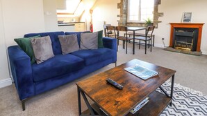 A living room with a blue sofa, wooden coffee table, stone fireplace, and a dining area with chairs and a table. - Mearbeck Apartment (Settle)