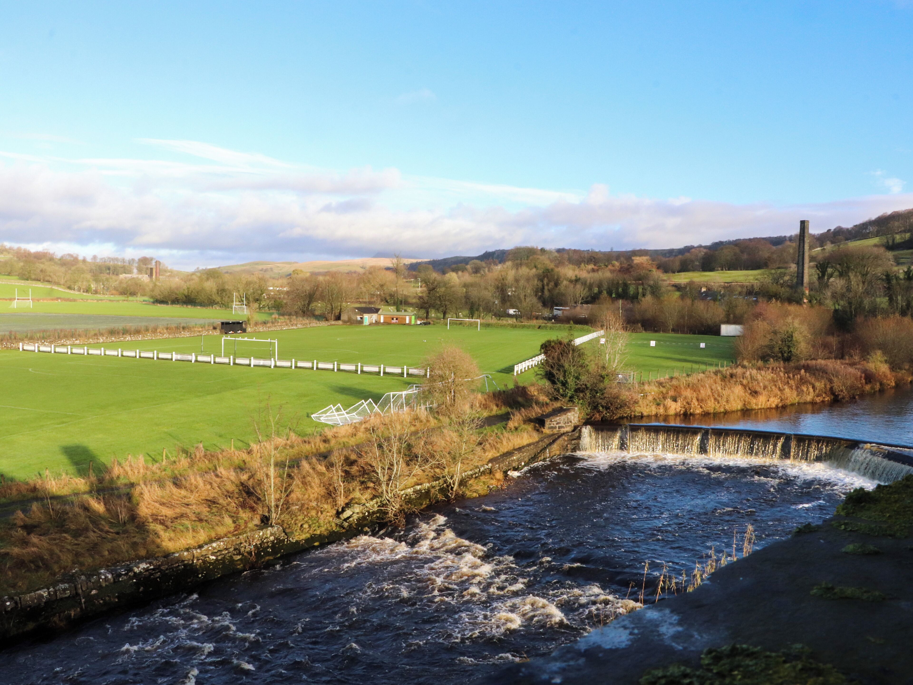 Une rivière traverse un champ verdoyant, avec un petit pont et, au loin, on aperçoit un terrain de football.