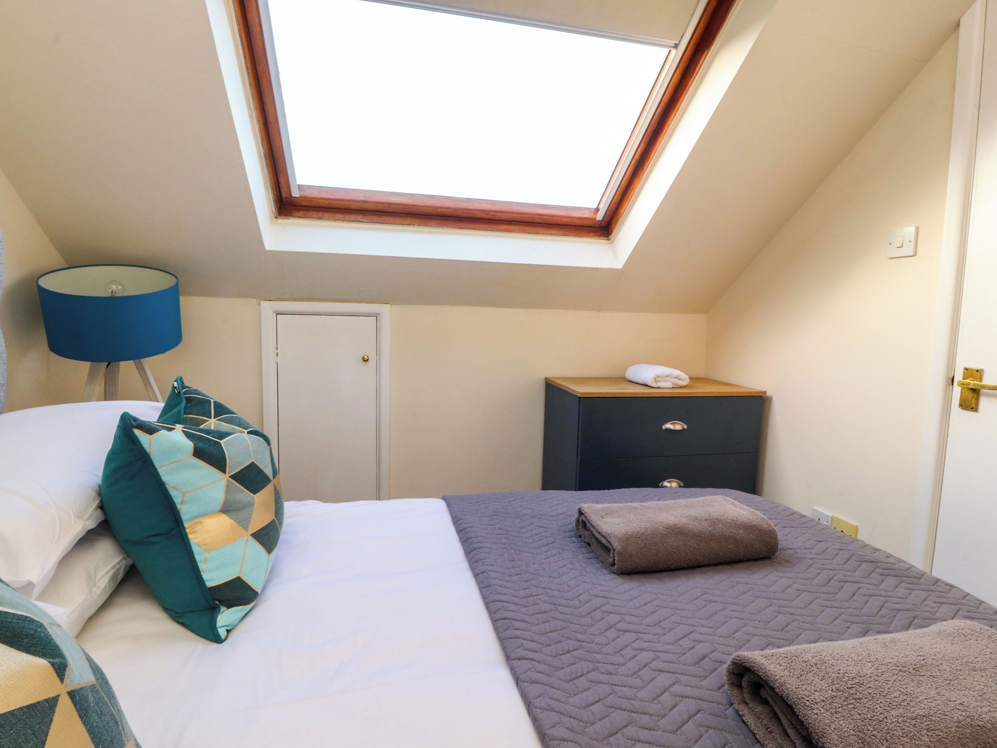 Une chambre avec un puits de lumière, un lit avec des oreillers, une table de chevet avec une lampe et une commode.