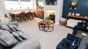 Una sala moderna con un sofá gris, una mesa de centro redonda y un sillón azul. Hay un comedor con una mesa y sillas de madera, y una cocina con estufa y armarios.