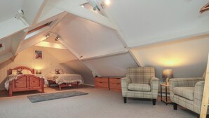Une chambre confortable située sous les toits, avec un plafond en pente, un lit en bois, deux fauteuils  à motifs écossais, une petite table avec une lampe et un tableau encadré accroché au mur.