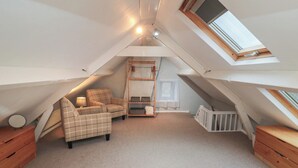 Une chambre mansardée chaleureuse avec un plafond haut, un puits de lumière, un fauteuil à carreaux, une petite table et un berceau.