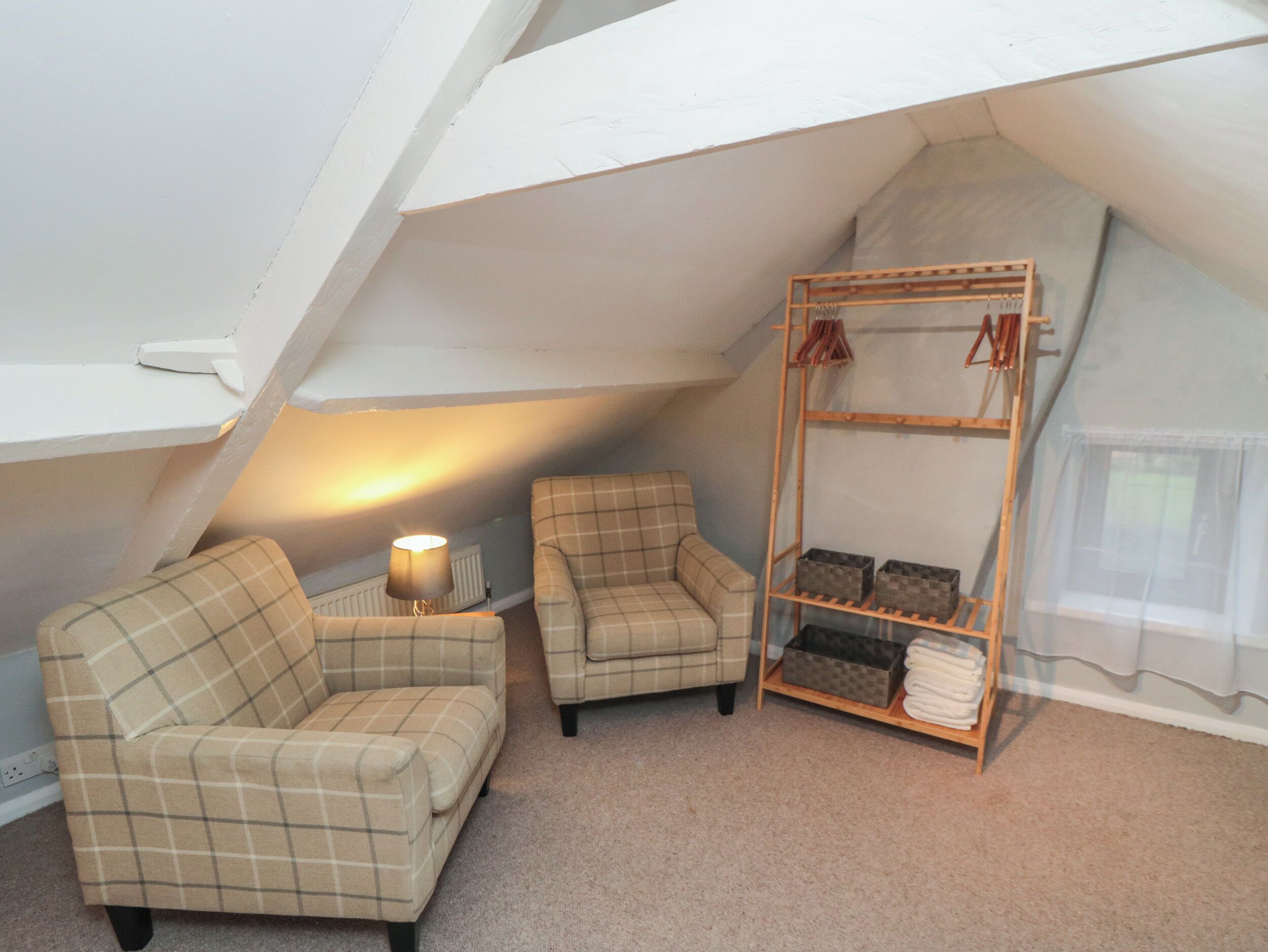 A cozy attic room with two plaid armchairs, a small table with a lamp, and a wooden shelf with storage boxes and folded towels.