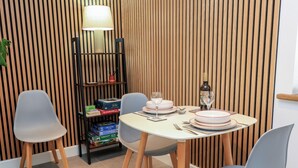 Ein Essbereich mit einem runden Tisch für zwei Personen, zwei grauen Stühlen, einer Wand aus Holzlatten, einem Regal mit Büchern und einer Lampe sowie einer Weinflasche.