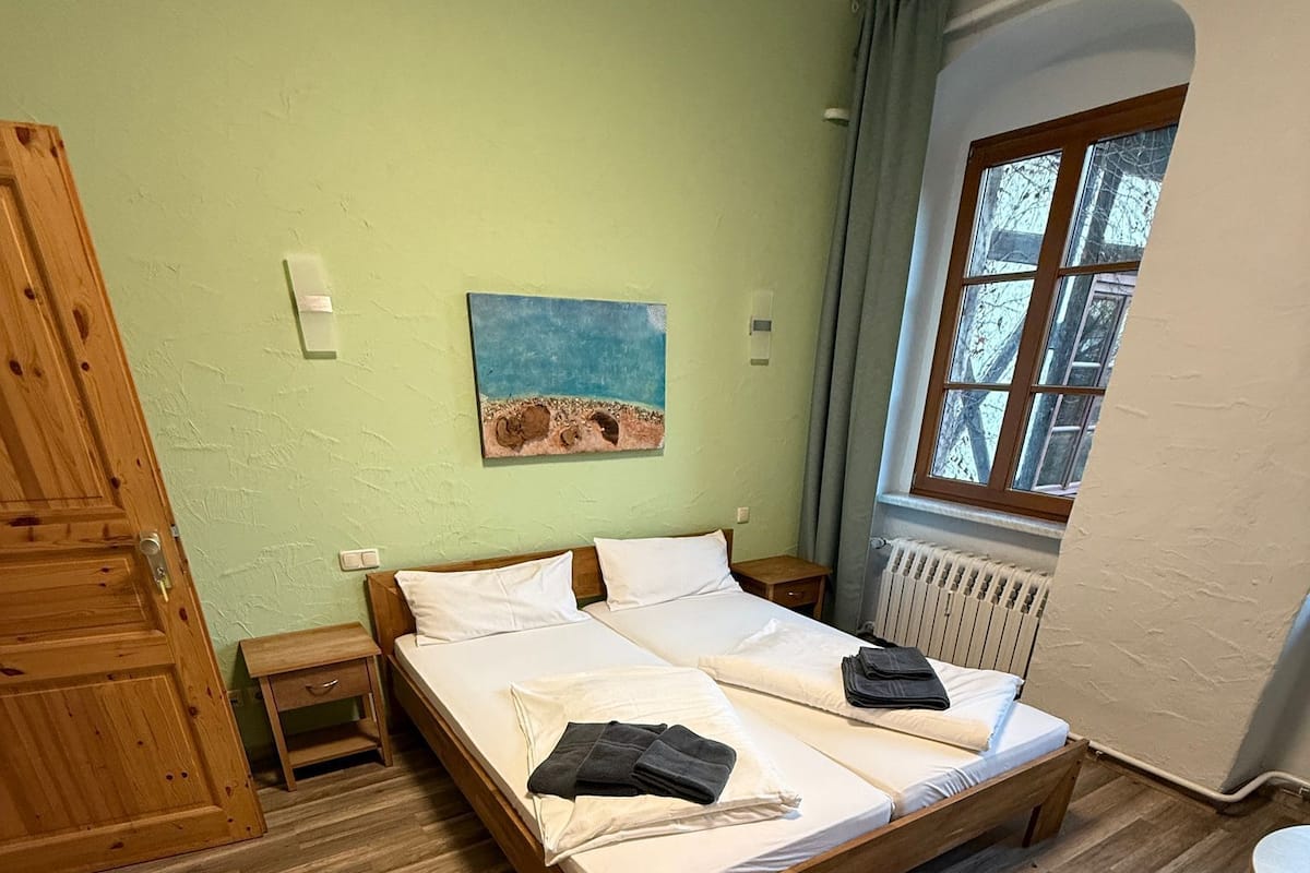 A room with a wooden bed, a small bedside table, a radiator, and a window with curtains.