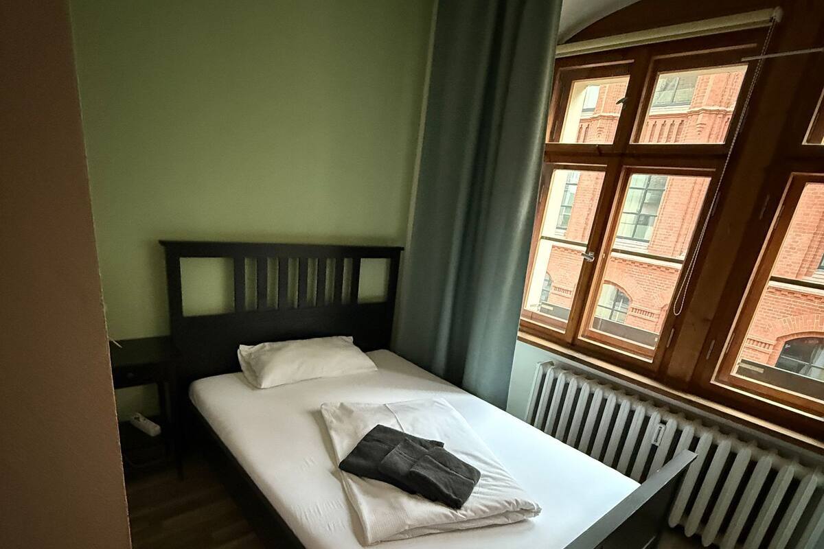 A single bed with a wooden headboard, white linens, and a folded towel. A window with a view of brick buildings is visible.