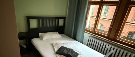 A single bed with a wooden headboard, white linens, and a folded towel. A window with a view of brick buildings is visible.