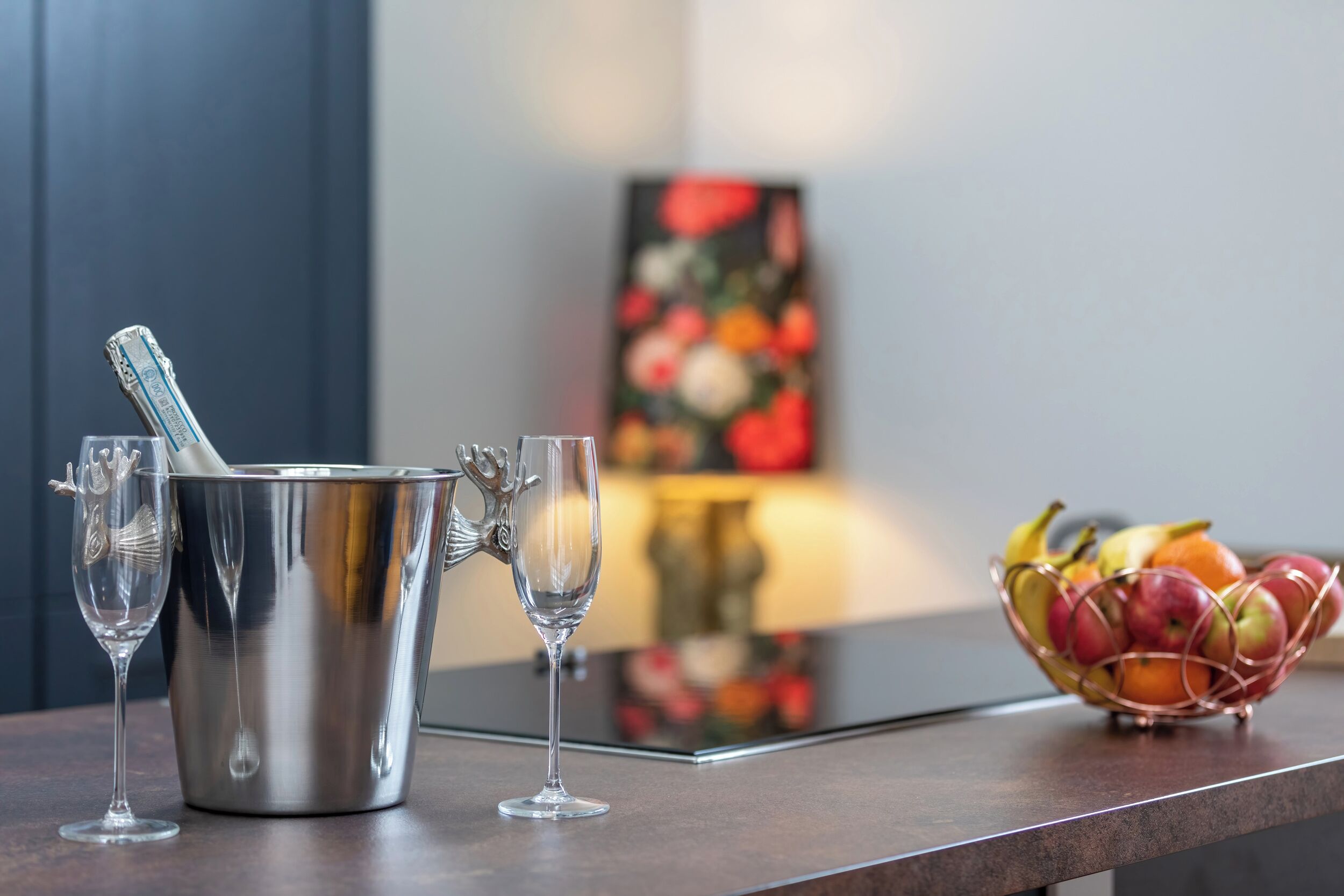 A metallic bucket with a spoon, two wine glasses, and a fruit basket on a reflective surface.