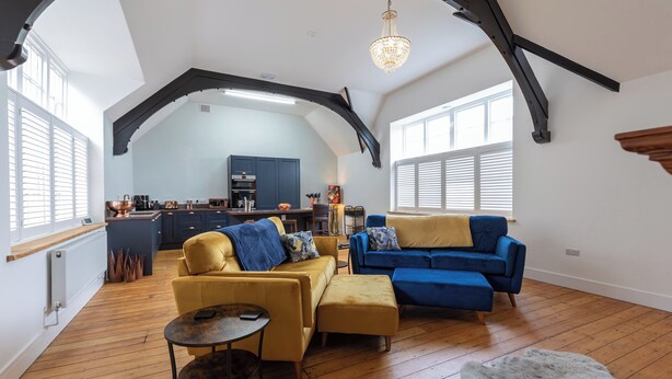 Een ruime loft met hoge plafonds, zichtbare balken en grote ramen. Er staan een gele en een blauwe bank, en een eethoek met een keuken op de achtergrond.