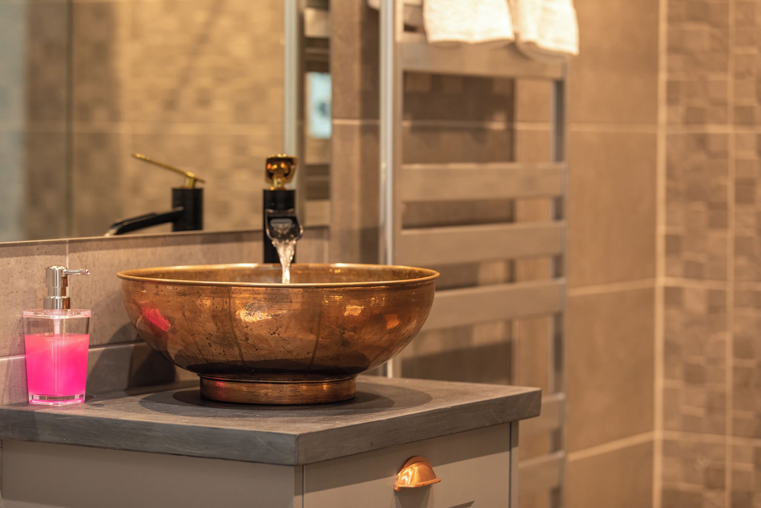 A bathroom with a copper sink, a glass soap dispenser, and a mirror reflecting a towel rack.