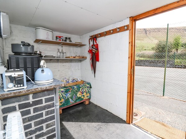 A compact kitchen with a toaster, kettle, and a small refrigerator. There is a wooden bench with a colorful cushion, and a door leading to an outdoor area with a fence.