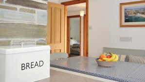 Une cuisine avec un mur en pierre, un garde-pain, un panier de fruits et un lit visible à travers une porte ouverte.