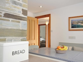 Una cocina con pared de piedra, un panero, un frutero y una cama visible a través de una puerta abierta.