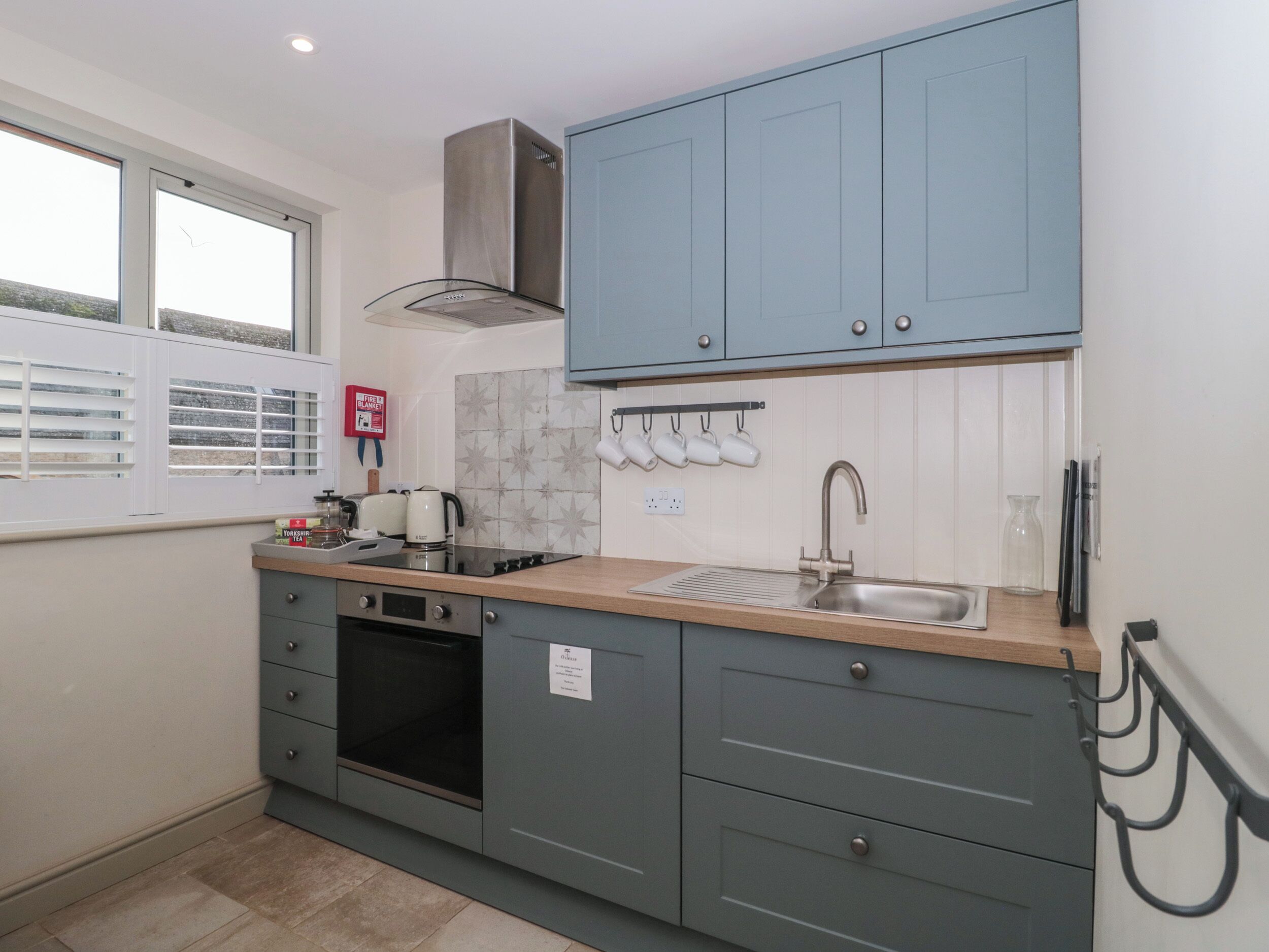A compact kitchen with grey cabinets, a stainless steel range hood, a built-in oven, a sink, and a window with blinds.