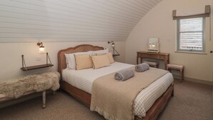 Een slaapkamer met een schuine plafond, een houten bed met geruit beddengoed, een bankje met een bont plaid, een kaptafel met spiegel en een raam met zonwering.