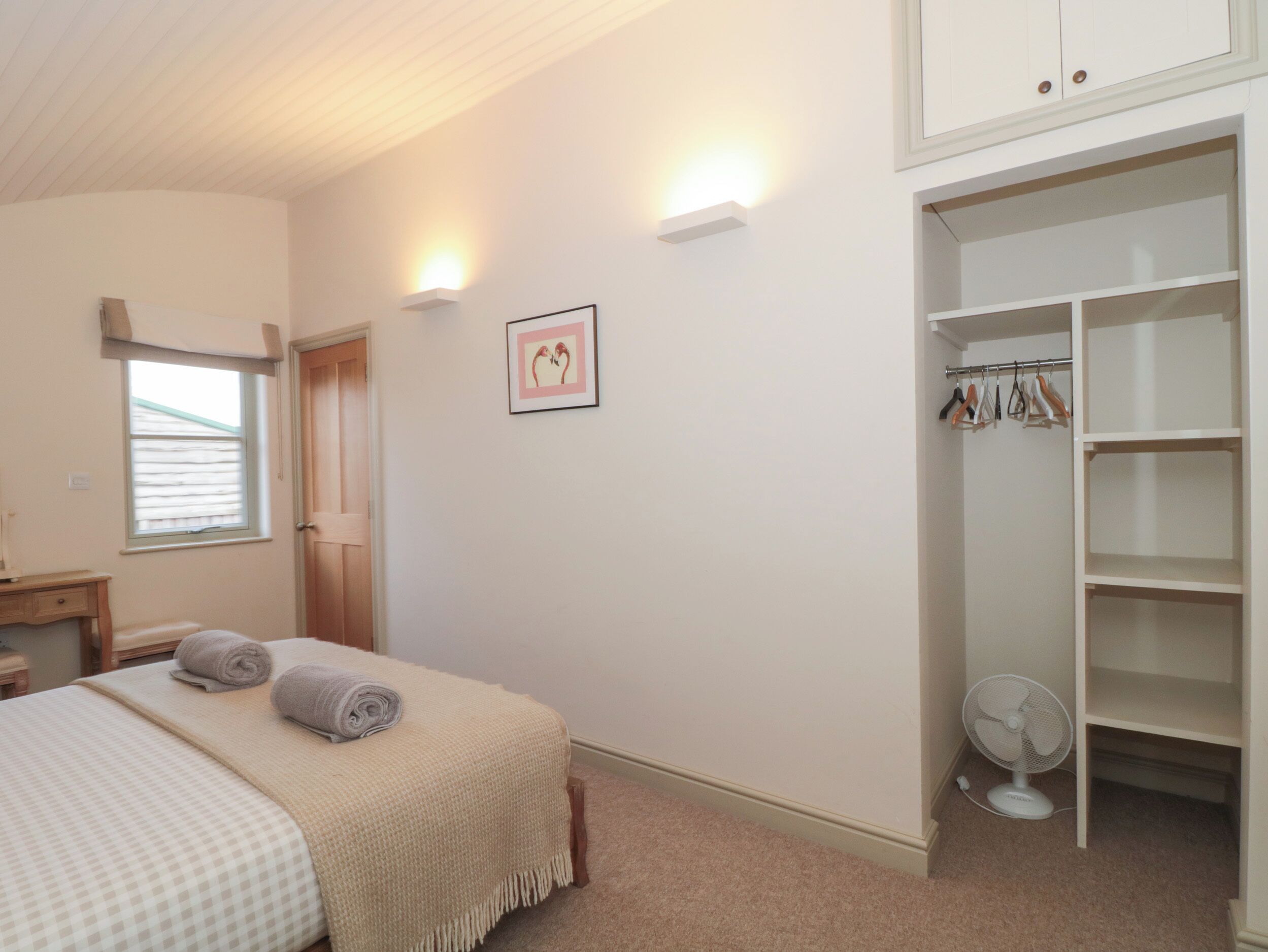 A bedroom with a bed, a bedside table, a wardrobe, a wall-mounted fan, and a window with blinds.