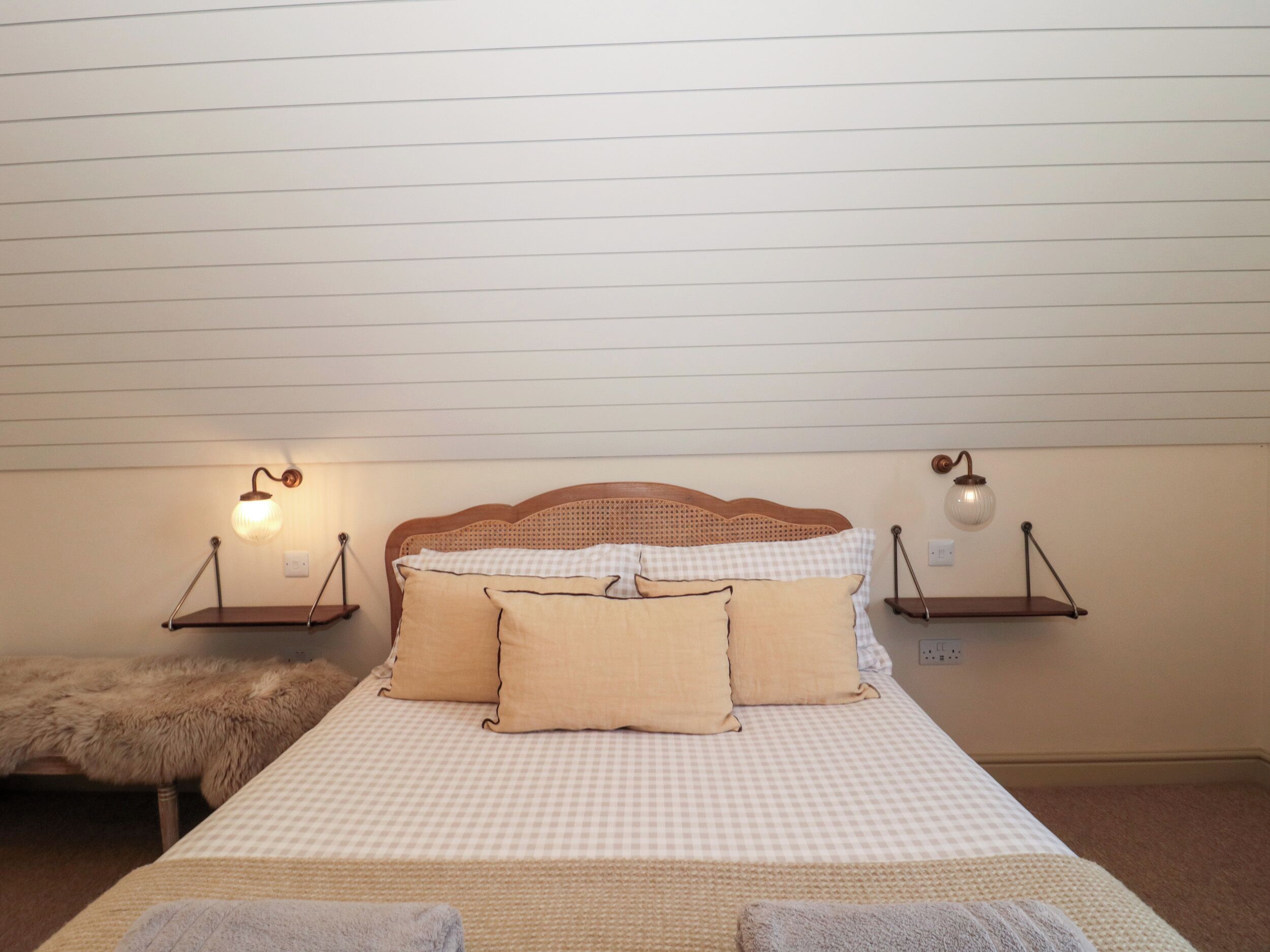 A neatly made bed with checkered bedding, two bedside tables with lamps, a fluffy bench, and a wooden headboard.