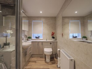 A modern bathroom with a toilet, sink, and mirror. There are two windows with blinds, and a radiator is visible on the right. - Bayside Cottage (Forres)