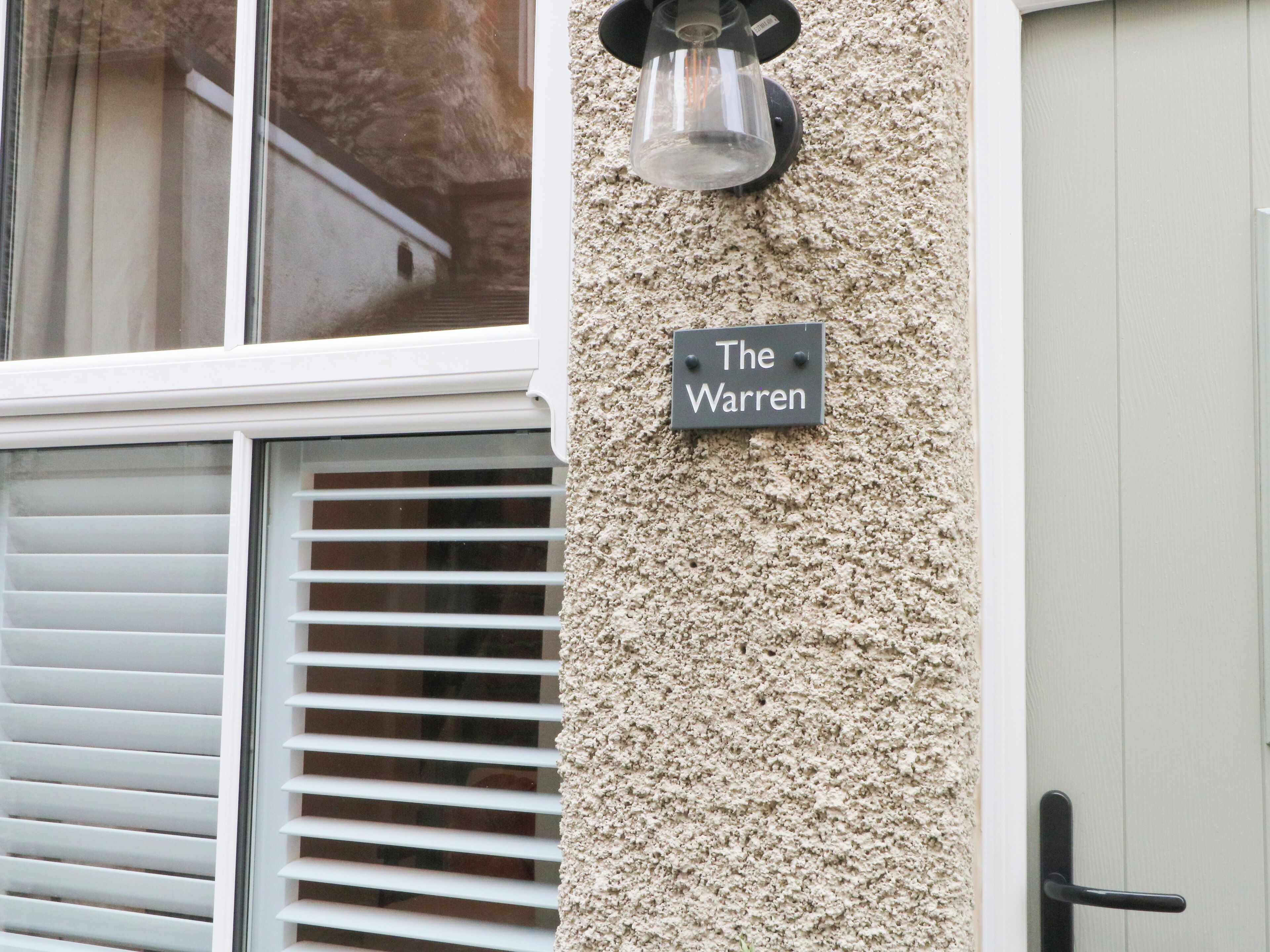 Uma placa com o número da casa numa parede texturizada, uma janela com persianas e uma porta.