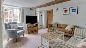 A living room with a television on a wooden stand, a grey armchair, a brown sofa, and a coffee table with a vase of flowers. - Amy Cottage (Wareham)