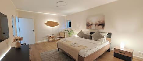 Un dormitorio moderno con una cama grande, mesitas de noche, un escritorio, una silla, una planta y un cuadro en la pared.