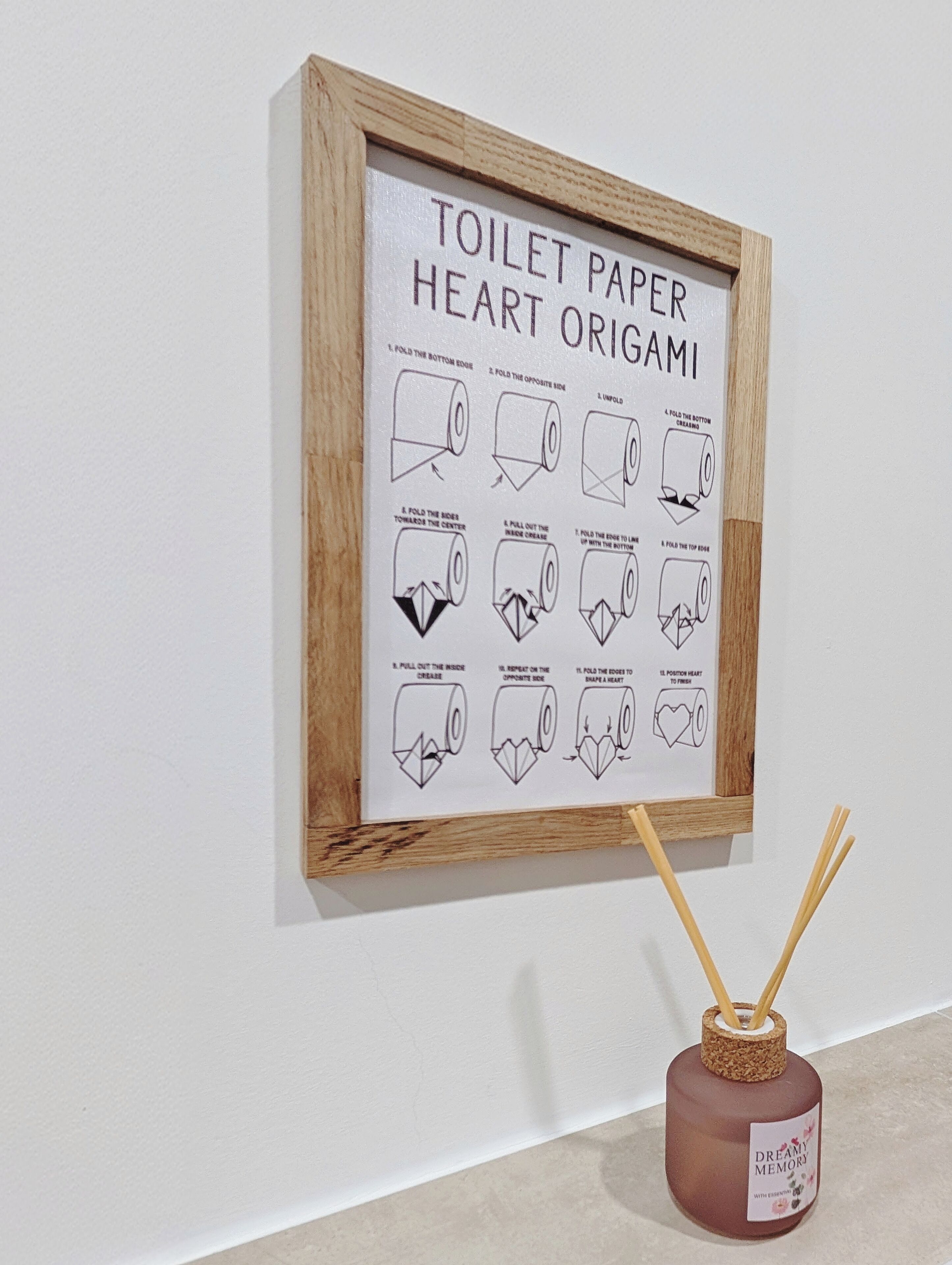 A framed origami heart made from toilet paper instructions on a wall, with a reed diffuser nearby.