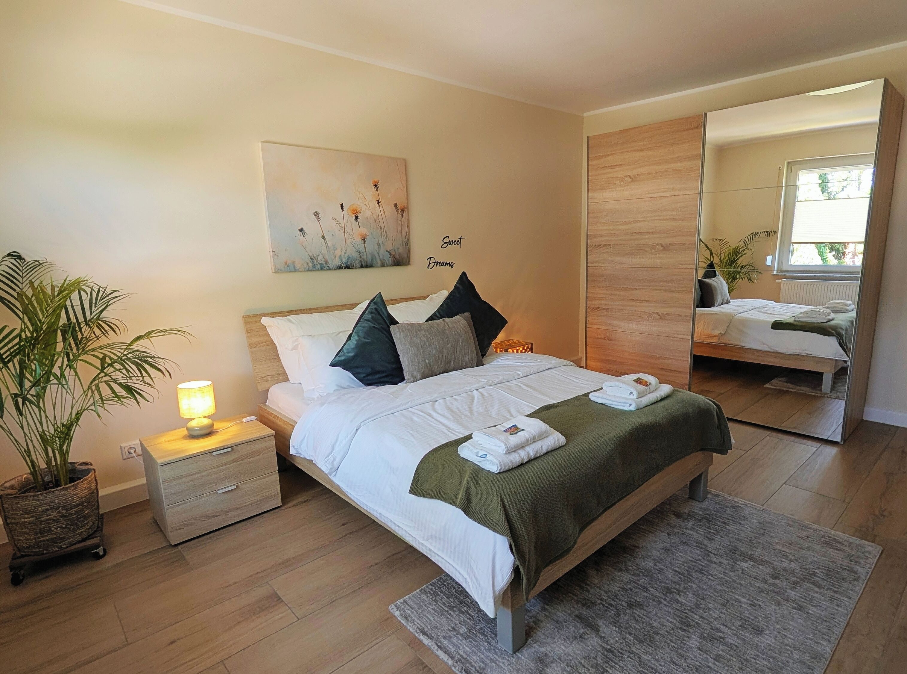 Quarto com cama, mesa de cabeceira com luminária, planta em vaso, obra de arte e espelho.
