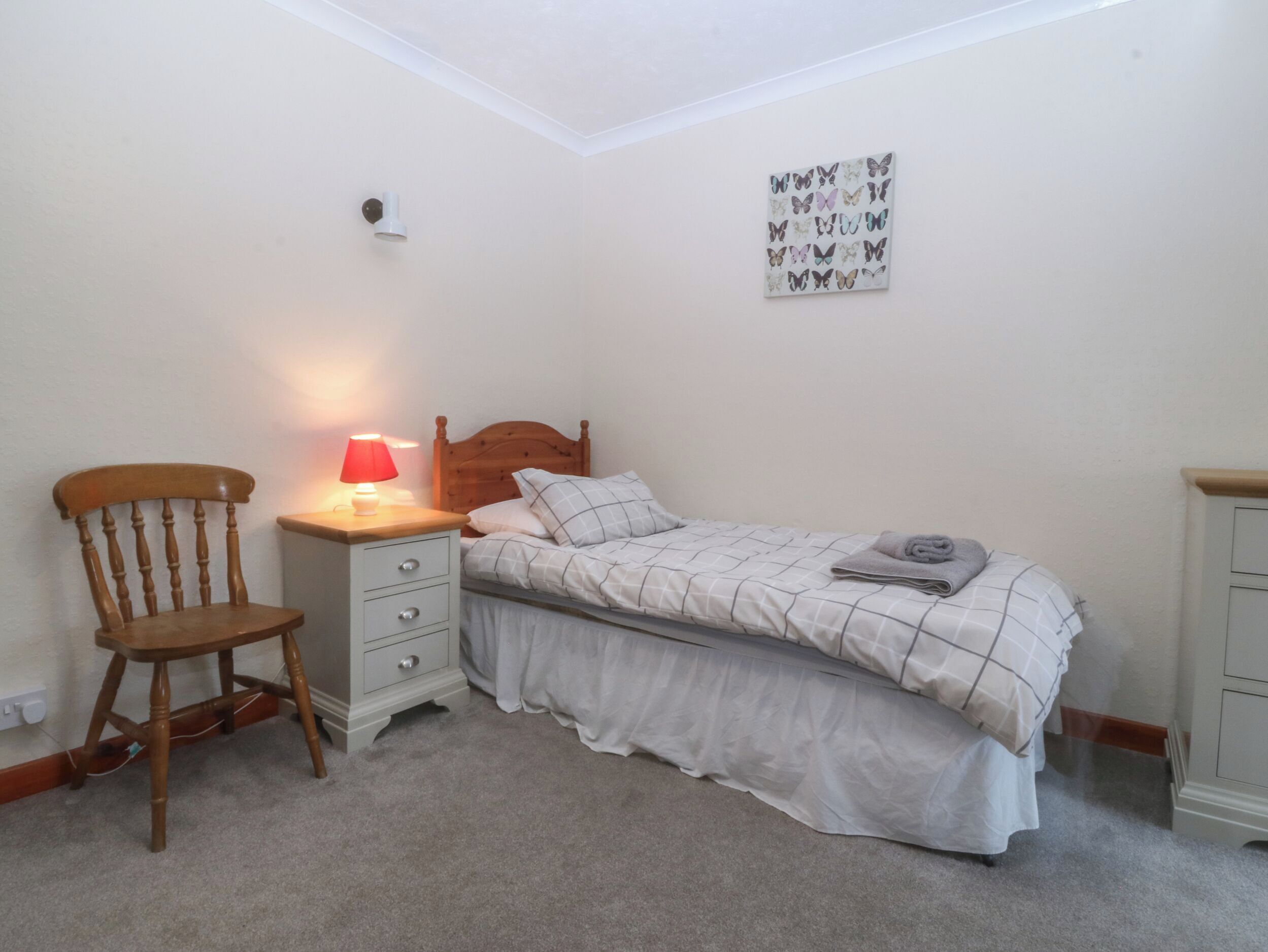 A bedroom with a bed, a chair, a bedside table with a lamp, a wall-mounted artwork, and a carpeted floor.