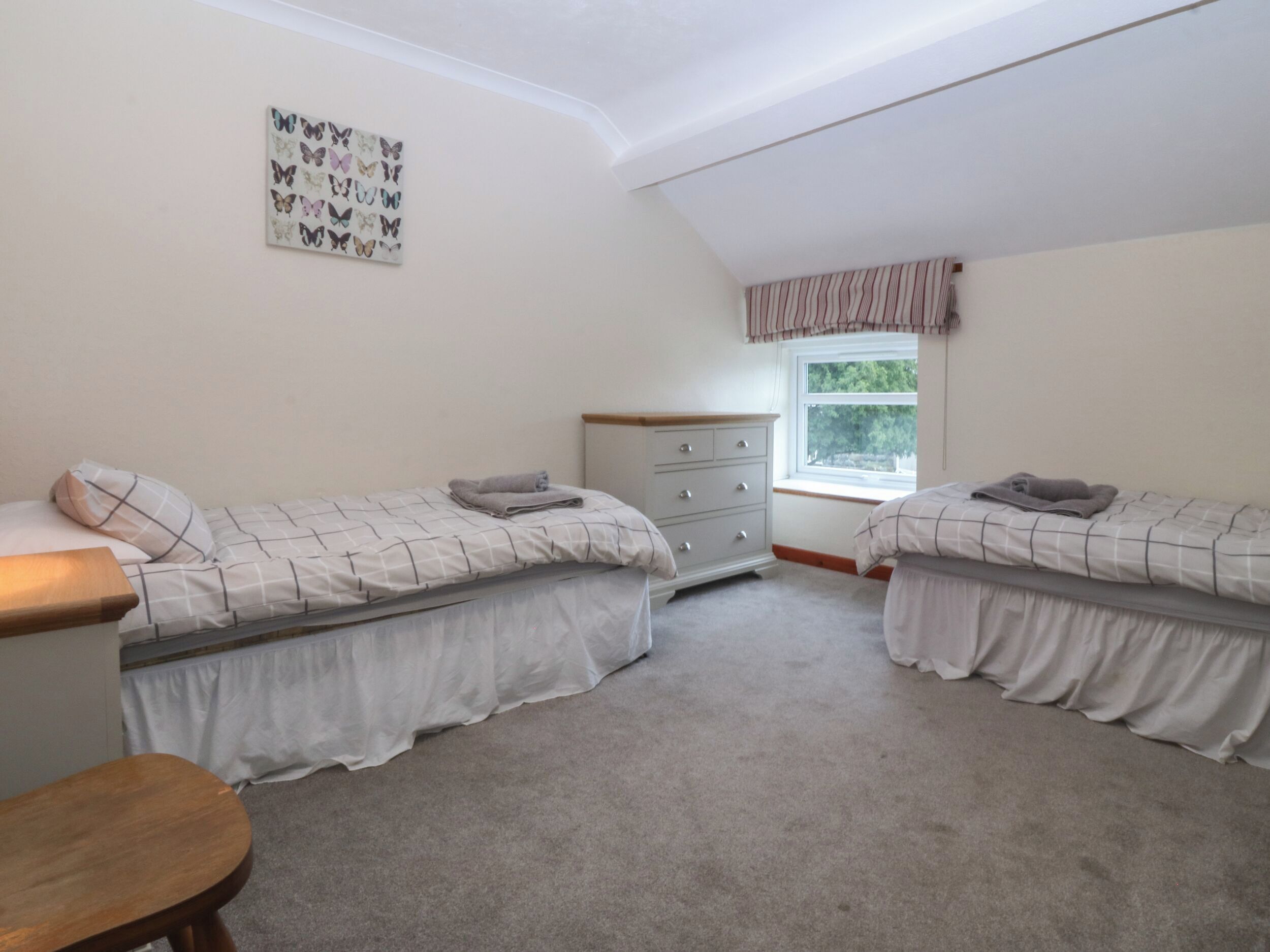 A bedroom with two single beds, a wooden nightstand, a dresser, and a window with a view of greenery.