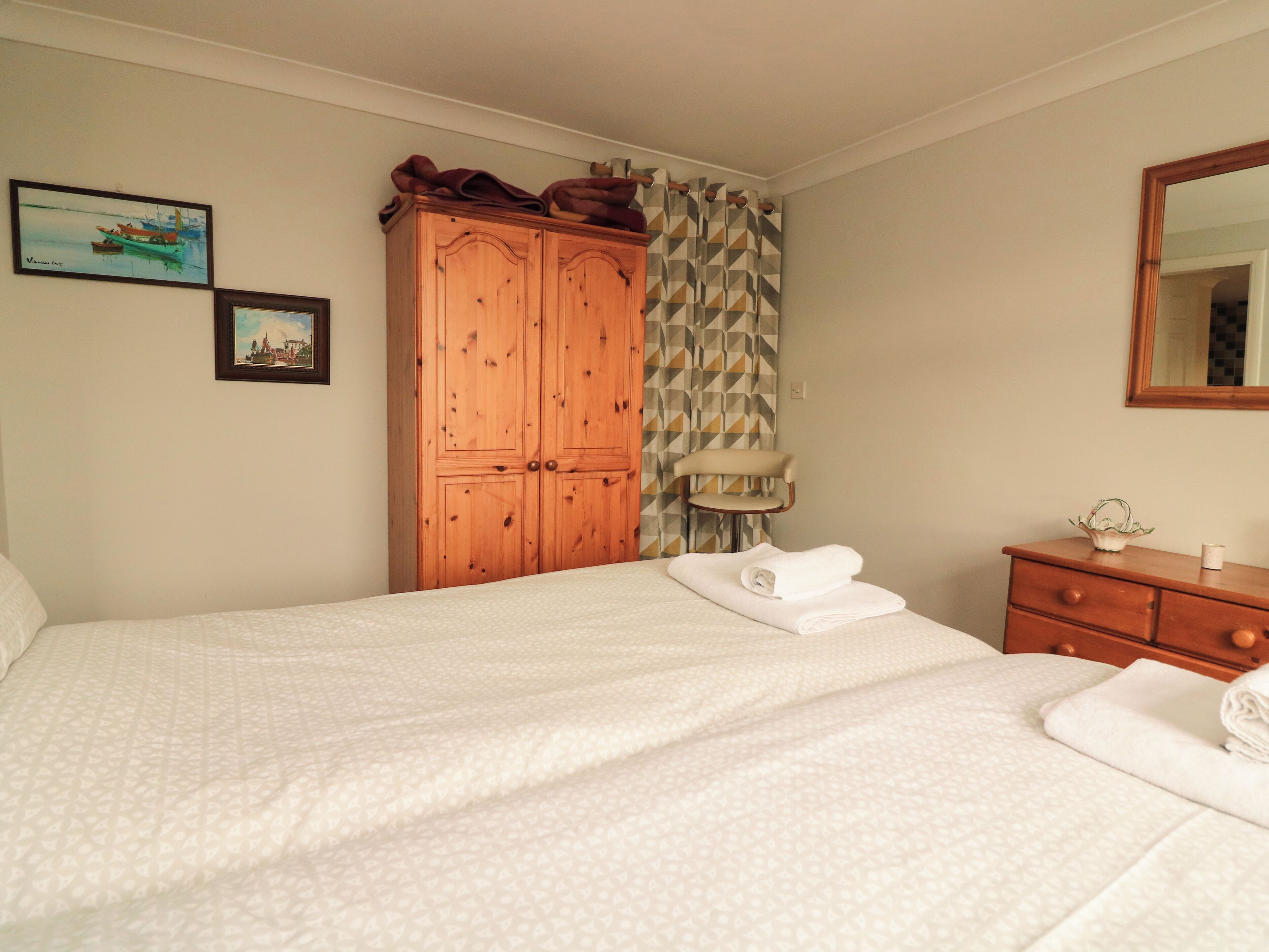 A neatly made bed with a patterned bedspread, a wooden wardrobe, a chair, and a mirror on the wall.