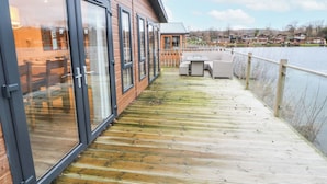 Une terrasse en bois avec une porte vitrée donnant sur une pièce, une table et des chaises, et une vue sur un lac avec des maisons en arrière-plan.