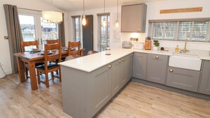 Une cuisine moderne avec des meubles gris, un plan de travail blanc et un îlot central. On y trouve un coin repas avec des chaises en bois et une table, et une vue sur l’extérieur grâce à de grandes fenêtres.