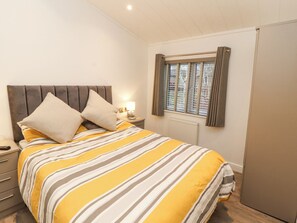 A bedroom with a bed, a nightstand, a lamp, a window with curtains, and a radiator. - 12A Lake Escape (Carnforth)
