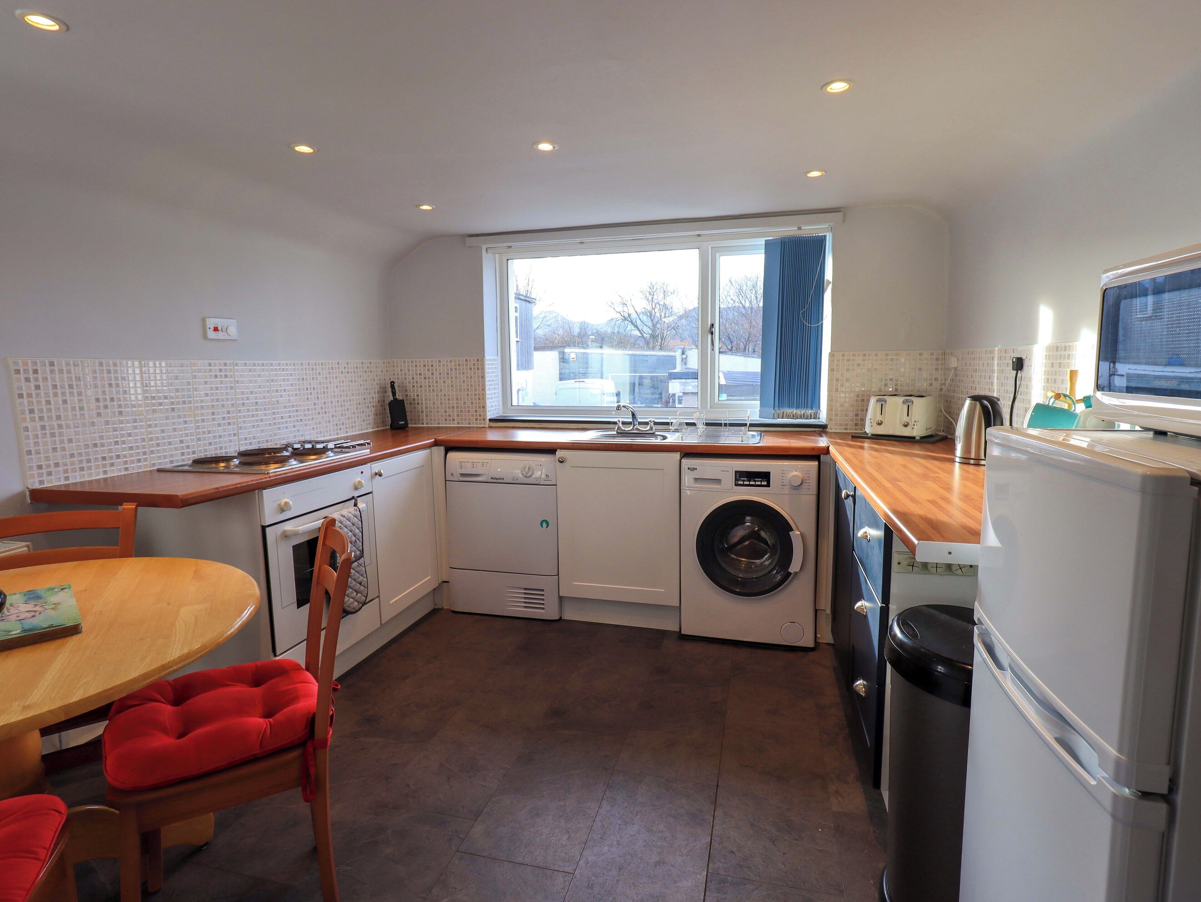 A compact kitchen with a dishwasher, washing machine, and a microwave. There is a dining table with chairs and a window providing natural light.