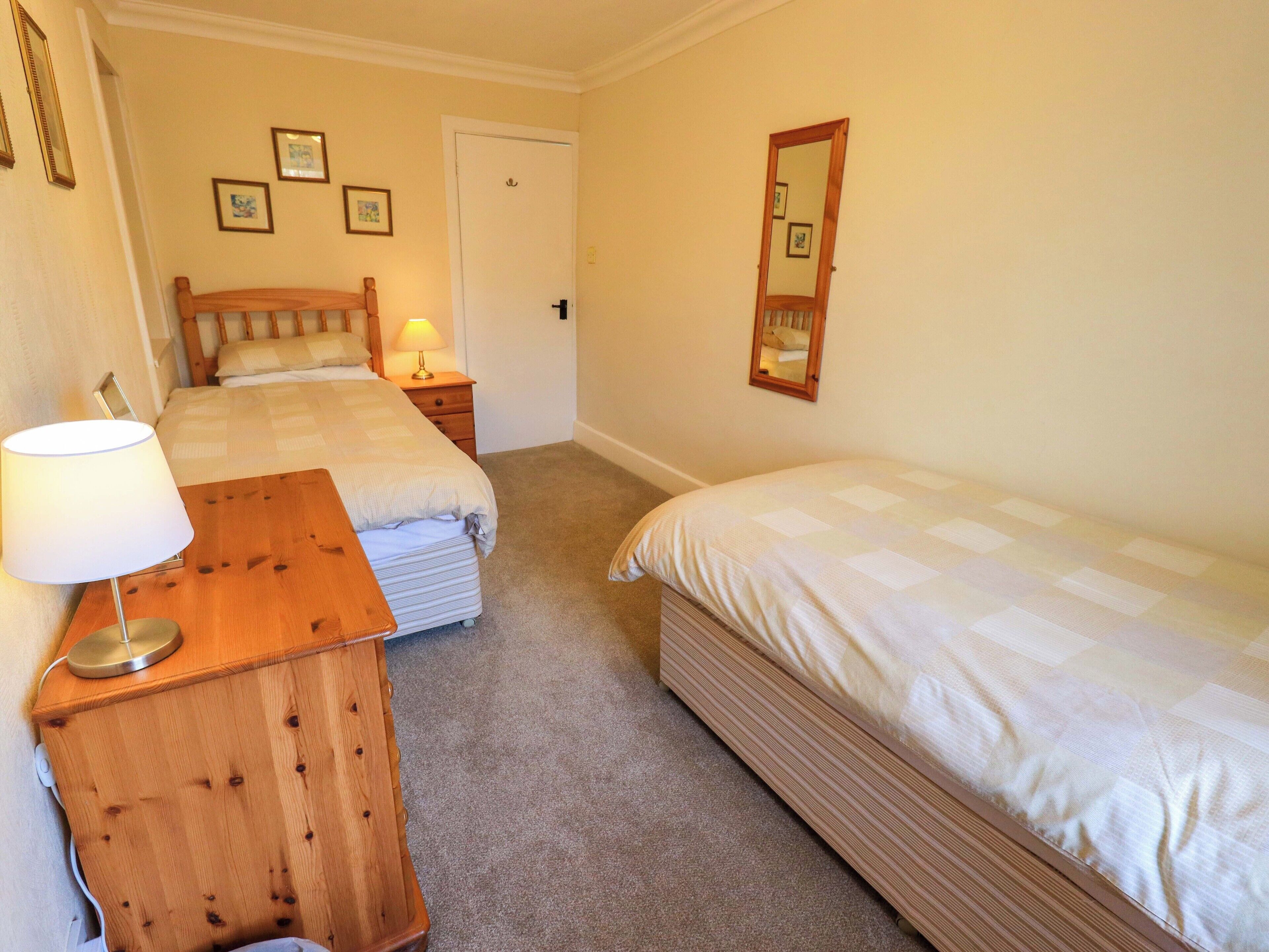 A bedroom with two beds, a bedside table with a lamp, a wooden dresser, and a door.