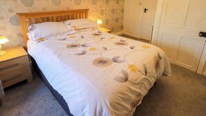 Un lit bien fait, avec une literie à motifs floraux, une tête de lit en bois, une table de chevet avec une lampe, et une porte visible au fond.