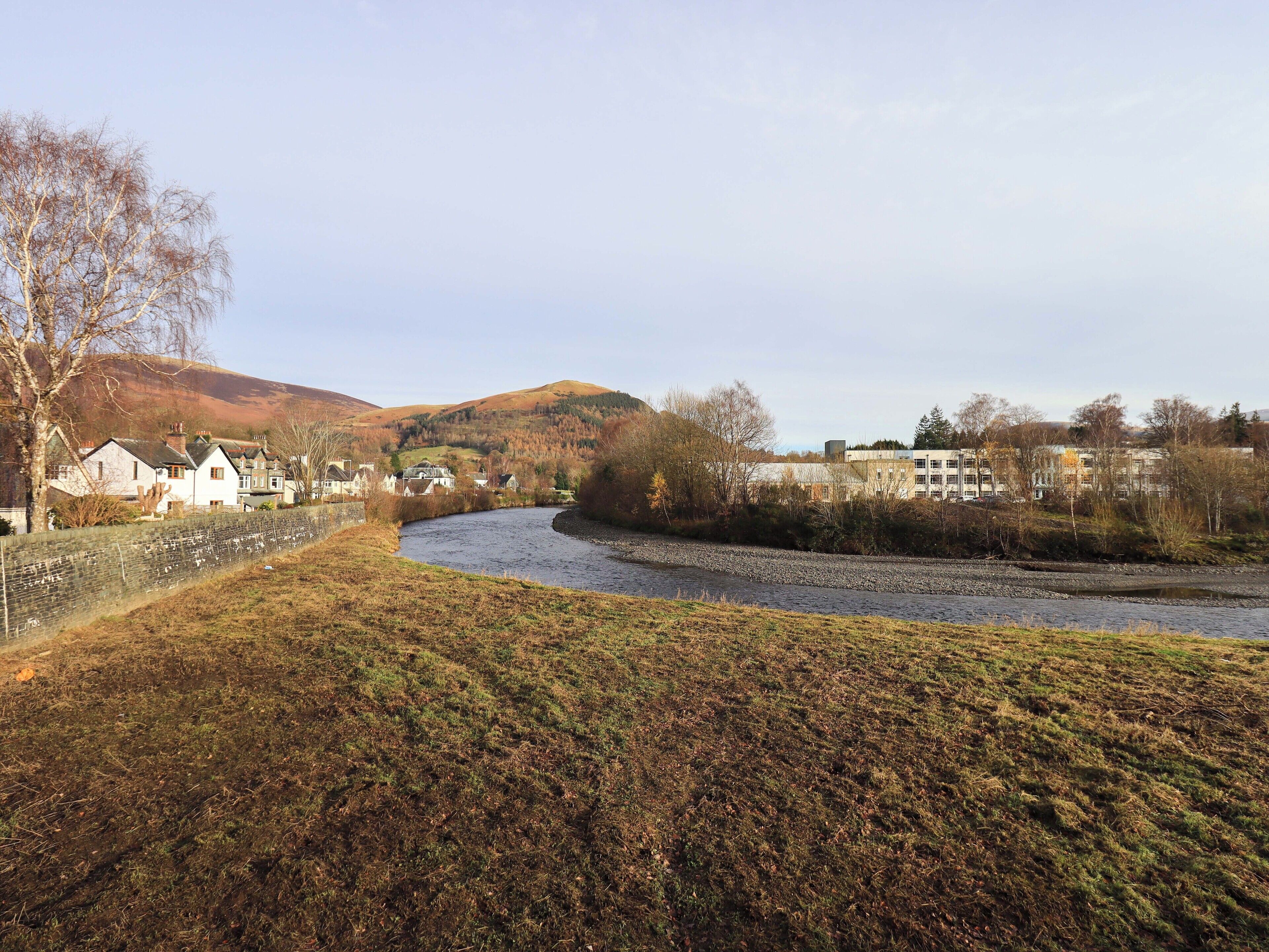 A river flowing through a rural area with houses on one side and a building complex on the other.