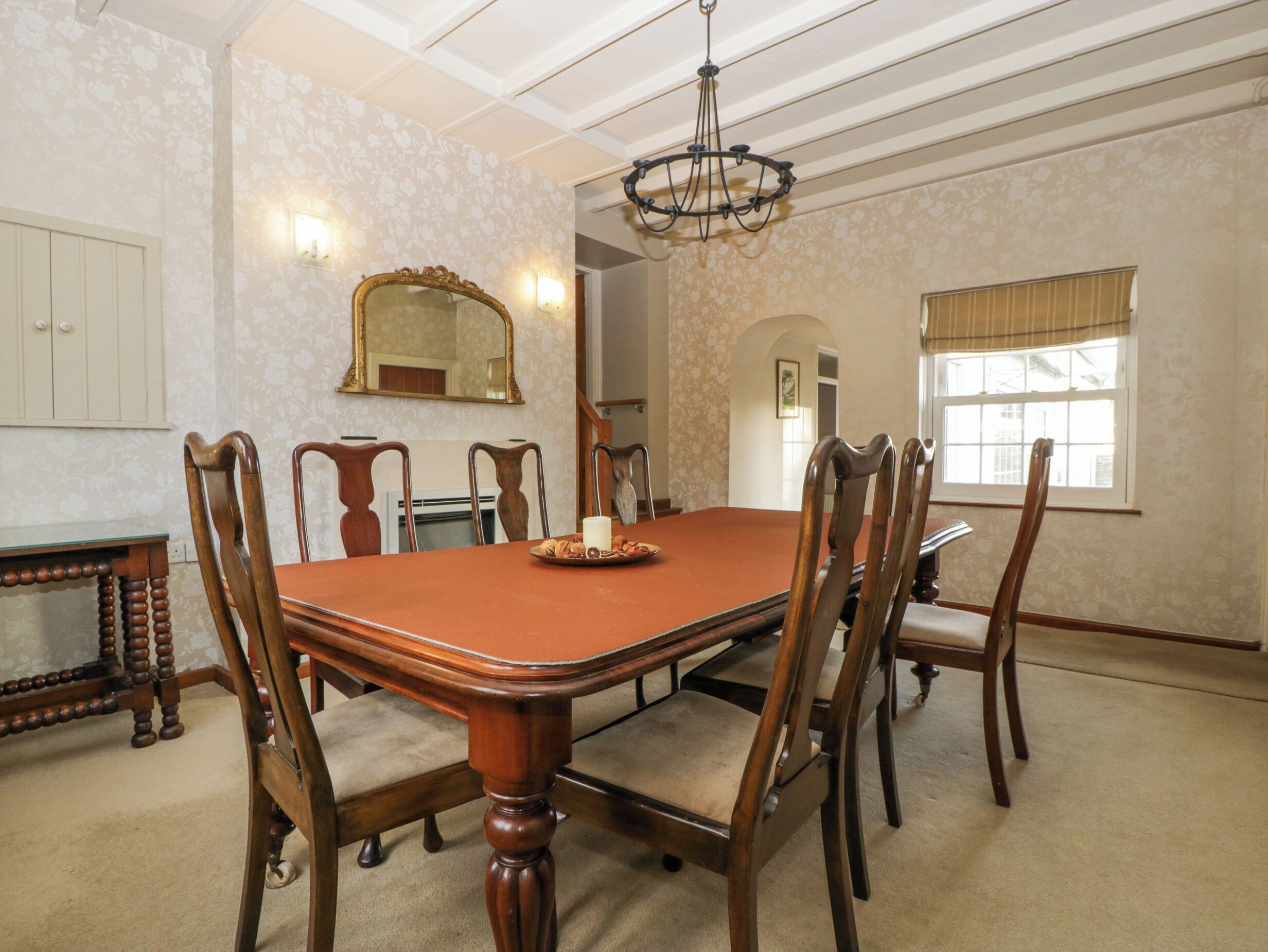 A dining room with a large wooden table, six chairs, a chandelier, and a window with curtains.