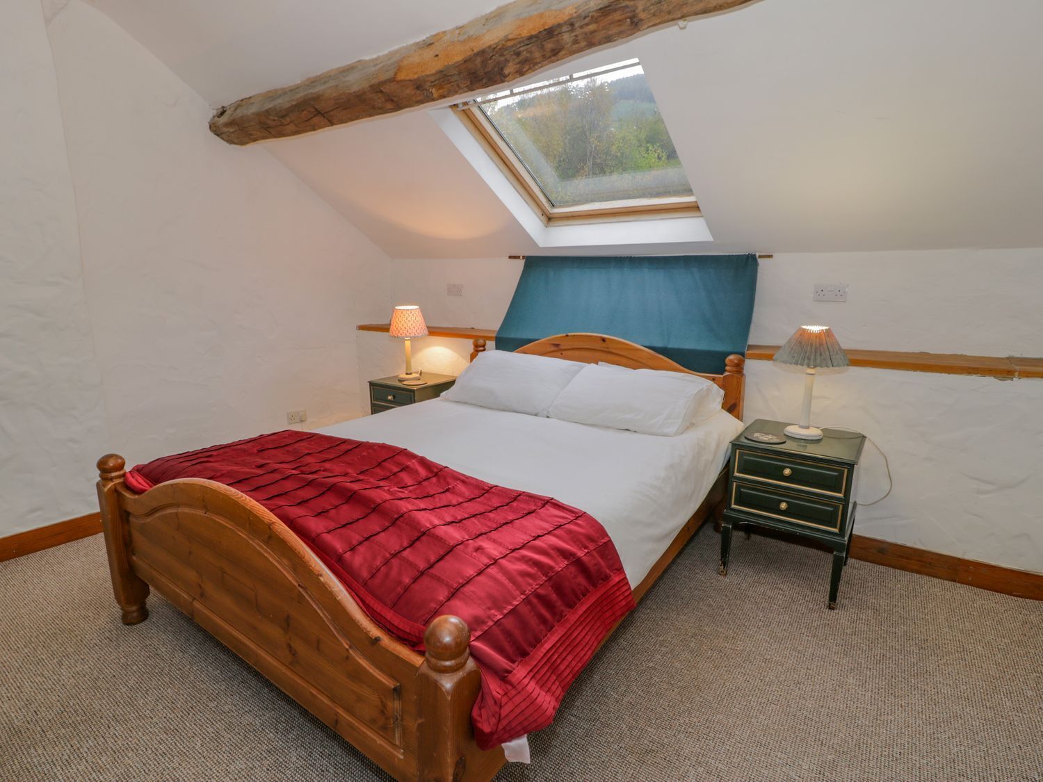 Ein Schlafzimmer mit geneigter Decke, einem Oberlicht, einem hölzernen Bett, zwei Nachttischen mit Lampen und einer roten Bettdecke.