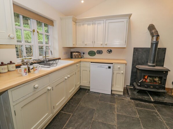 A kitchen with white cabinets, a dishwasher, a toaster, and a black wood-burning stove. - Ysgubor (Machynlleth)