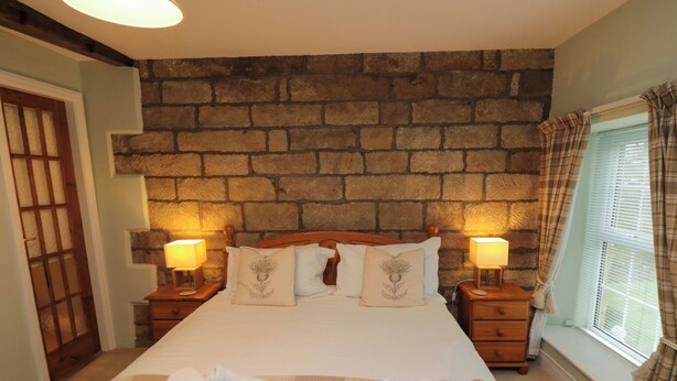 A neatly made bed with pillows, a checkered blanket, and rolled towels on a carpeted floor. There are two bedside tables with lamps, a stone wall behind the bed, and a window with curtains. - The Old Post Office (Pickering)
