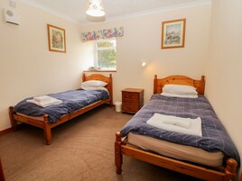 Una habitación con dos camas de madera, cada una con colcha azul y almohadas blancas. Hay una mesita de noche de madera entre las camas, una ventana con cortina floral y un aire acondicionado instalado en la pared.