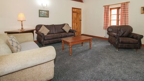 A living room with a brown leather sofa, a wooden coffee table, and a brown leather armchair. There is a wooden door and a window with plaid curtains. - Walton Maisonette (Craven Arms)