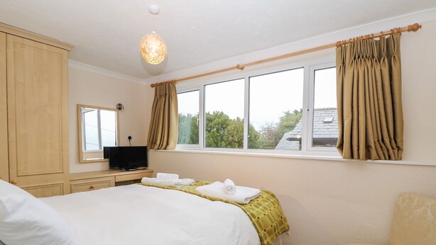 A bedroom with a bed, a television, a window with curtains, a chair, and a hanging light fixture. - Y Bwthyn (Pwllheli)