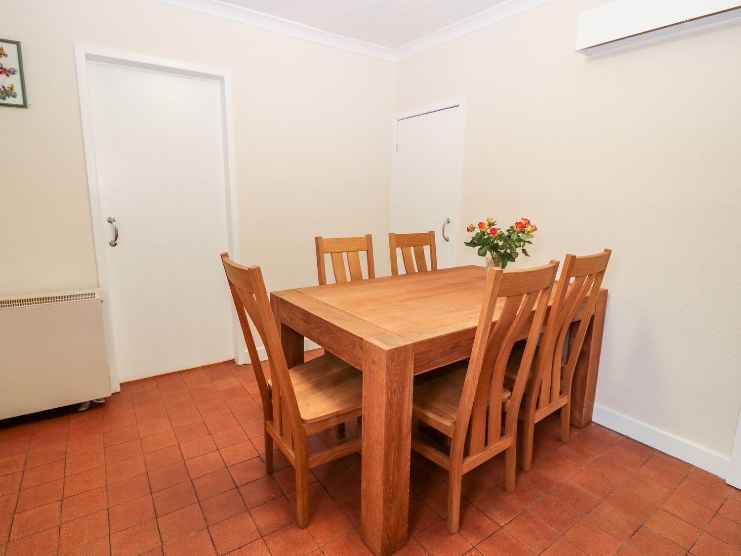 Un comedor con mesa de madera y seis sillas, un pequeño jarrón con flores y un refrigerador blanco.
