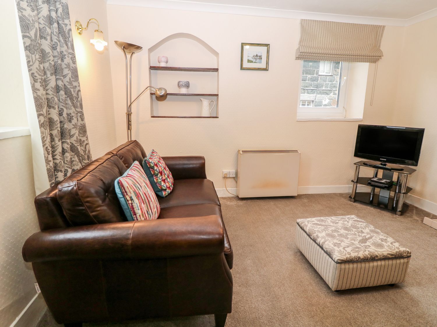 A living room with a brown leather sofa, a patterned ottoman, a TV on a stand, a radiator, and a window with a view.