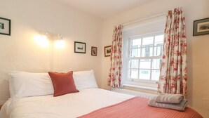 Een hotelkamer met een bed, kussens, een raam met bloemen gordijnen en een wandlamp.