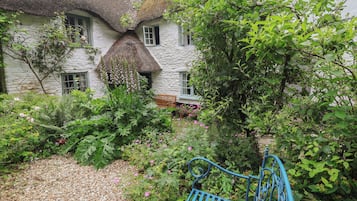 A rustic stone building with a thatched roof, surrounded by lush greenery and a gravel path.