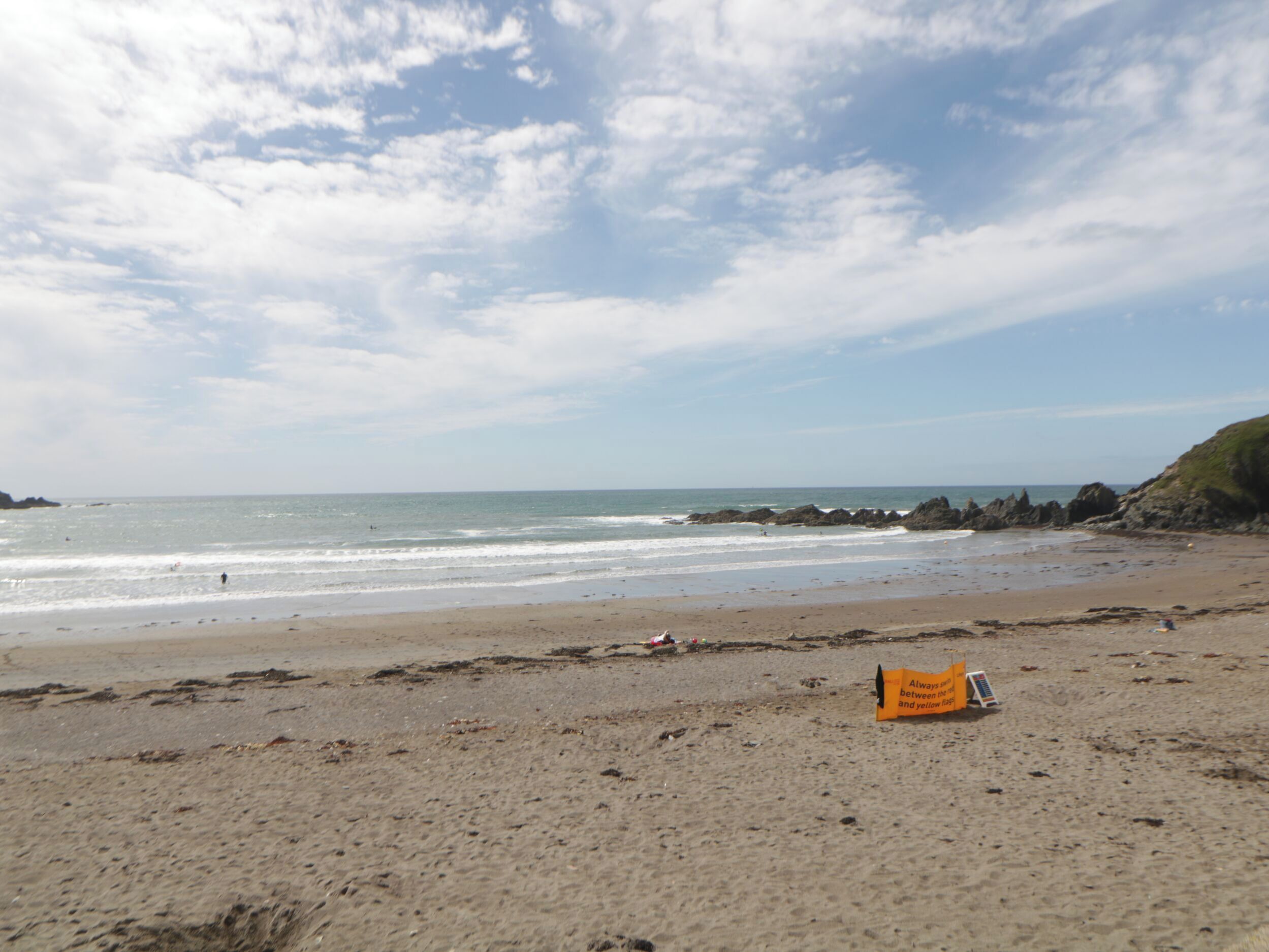 Een strand met een heldere lucht, een rotsachtige kustlijn en een geel bord in het zand.