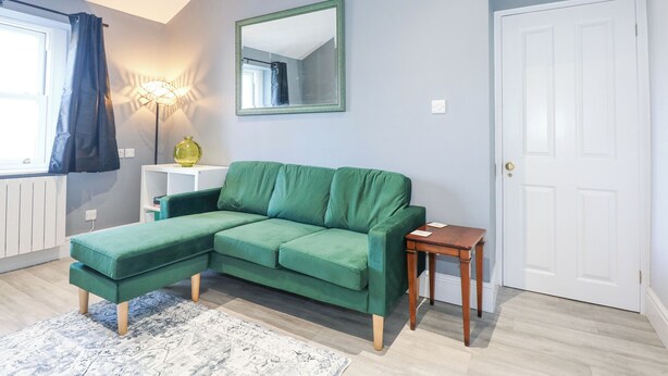 A modern living room with a green sectional sofa, a small wooden side table, a floor lamp, and a white door. - The Fir Grove Annex (Menai Bridge)